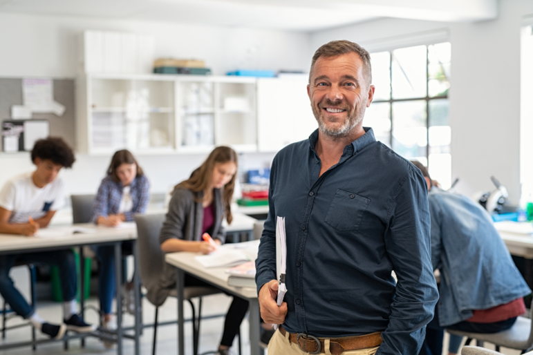 Lehrer steht im Klassenzimmer und lächelt in die Kamera; seine Schüler und Schülerinnen sitzen hinter ihm am Tisch und schreiben