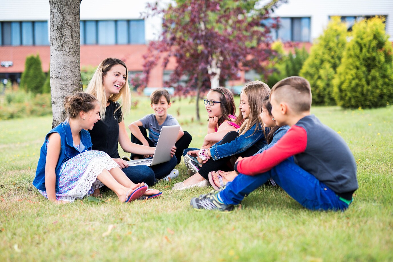 Eine Lehrerin sitzt mit fünf Kindern auf einer Wiese vor einem Schulgebäude und zeigt ihnen etwas auf einem Laptop.